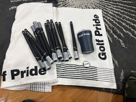 Thirteen Golf Pride grips laid out on two Golf Pride towels, with a ball marker and a tumbler.